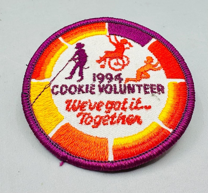 K&ouml;nnte beinhalten: Runder, gestickter Aufn&auml;her mit lila Rand und wei&szlig;er Mitte. Der Aufn&auml;her zeigt den Text "1994 Cookie Volunteer" und "We've got it... Together." Silhouetten von Personen sind in Orange und Lila dargestellt, mit einem Farbverlauf von Orange, Gelb und Lila.
