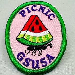 K&ouml;nnte beinhalten: Ovaler Aufn&auml;her mit rosa Hintergrund und gr&uuml;nem Rand. Die Worte "PICNIC" und "GSUSA" sind in Lila gehalten. Eine Wassermelonen-Scheibe wird von einer schwarzen Ameise getragen. Die Wassermelone ist rot, wei&szlig; und gr&uuml;n.