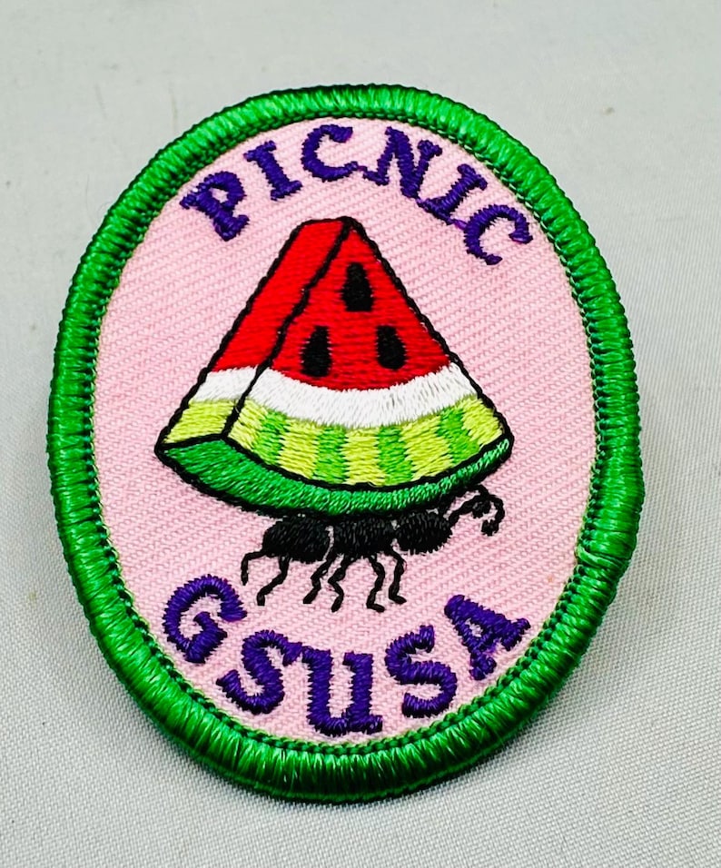 K&ouml;nnte beinhalten: Ovaler Aufn&auml;her mit rosa Hintergrund und gr&uuml;nem Rand. Die Worte "PICNIC" und "GSUSA" sind in Lila gehalten. Eine Wassermelonen-Scheibe wird von einer schwarzen Ameise getragen. Die Wassermelone ist rot, wei&szlig; und gr&uuml;n.
