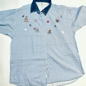 May include: Blue and white striped button-down shirt with a denim collar. The shirt features embroidered teddy bears, stars, and patriotic symbols. The shirt has short sleeves and a pocket on the left side.