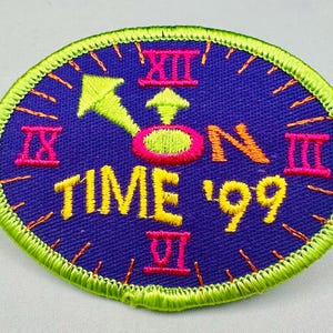 K&ouml;nnte beinhalten: Ovaler Aufn&auml;her mit einem Zifferblatt-Design. Der Aufn&auml;her hat einen dunkelblauen Hintergrund mit gelbem Text "TIME '99". Die Zeiger und Ziffern der Uhr sind neongr&uuml;n und pink. Der Rand ist neongr&uuml;n.