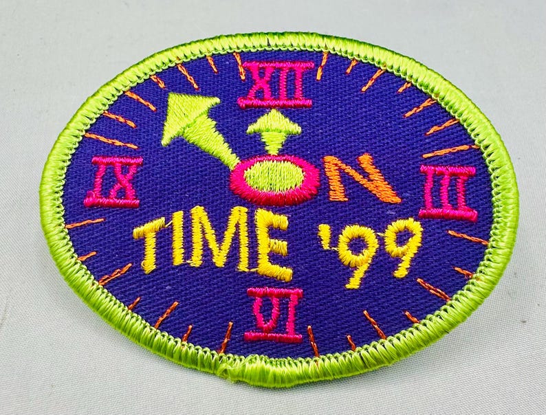 K&ouml;nnte beinhalten: Ovaler Aufn&auml;her mit einem Zifferblatt-Design. Der Aufn&auml;her hat einen dunkelblauen Hintergrund mit gelbem Text "TIME '99". Die Zeiger und Ziffern der Uhr sind neongr&uuml;n und pink. Der Rand ist neongr&uuml;n.