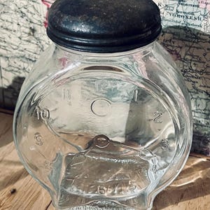 Puede incluir: Un tarro de cristal transparente con tapa de metal oscuro. El tarro tiene forma de esfera de reloj, con números en los bordes y manecillas. El tarro está sobre una superficie de madera.