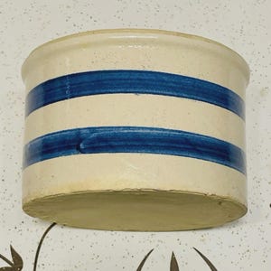 Puede incluir: Un tarro de gres vintage con base color crema y dos franjas azules. El recipiente cilíndrico tiene un borde ligeramente redondeado y una base sin esmaltar. Esta pieza rústica evoca la artesanía tradicional.