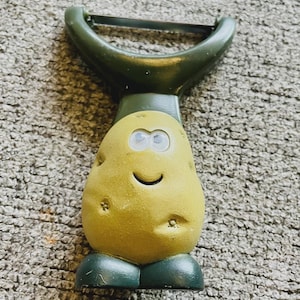 May include: A novelty potato peeler with a smiling potato-shaped handle. The peeler has a green handle and base, with a yellow potato body featuring eyes and a curved mouth. The blade is silver.