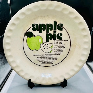 Può includere: Piatto da torta in ceramica bianco sporco con bordo smerlato. Il piatto presenta un'illustrazione di mela verde e le parole "apple pie" in un design circolare. Sono stampate anche le istruzioni della ricetta.