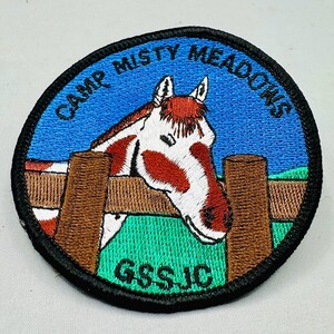 K&ouml;nnte beinhalten: Gestickter Aufn&auml;her mit einem Pferdekopf-Design. Der Aufn&auml;her zeigt ein Pferd mit einem rot-wei&szlig;en Muster, das &uuml;ber einen braunen Zaun blickt. Der Text "CAMP MISTY MEADOWS" ist oben in einem Bogen angeordnet, mit "GSS JC" unten.