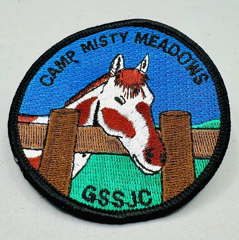 K&ouml;nnte beinhalten: Gestickter Aufn&auml;her mit einem Pferdekopf-Design. Der Aufn&auml;her zeigt ein Pferd mit einem rot-wei&szlig;en Muster, das &uuml;ber einen braunen Zaun blickt. Der Text "CAMP MISTY MEADOWS" ist oben in einem Bogen angeordnet, mit "GSS JC" unten.