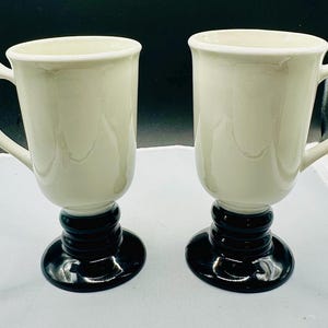 May include: Two cream-colored coffee mugs with a black base and handle. The mugs have a classic design, with a rounded top and a sturdy handle. The base is a dark, glossy black, and the mugs are clean and well-maintained.