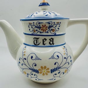 Puede incluir: Tetera de cerámica blanca con tapa, boquilla y asa. La tetera está decorada con motivos florales azules y dorados y la palabra "Tea" en letras negras. La tapa tiene un pequeño pomo.