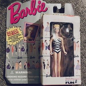 May include: A Barbie doll keychain in its original packaging. The doll is wearing a black and white striped swimsuit. The packaging includes the Barbie logo, the words "REALLY WORKS!" and a warning about choking hazards.