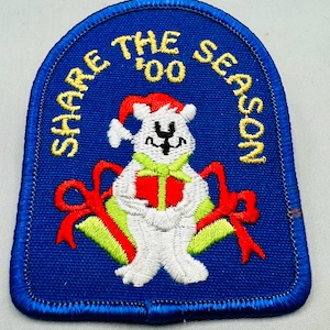 K&ouml;nnte beinhalten: Gestickter Aufn&auml;her mit blauem Hintergrund und einem wei&szlig;en B&auml;ren mit Weihnachtsm&uuml;tze. Der B&auml;r h&auml;lt ein gr&uuml;nes und rotes Geschenk. Der Text "SHARE THE SEASON '00" ist in Gelb gehalten. Rote Schleifen befinden sich auf beiden Seiten des B&auml;ren.