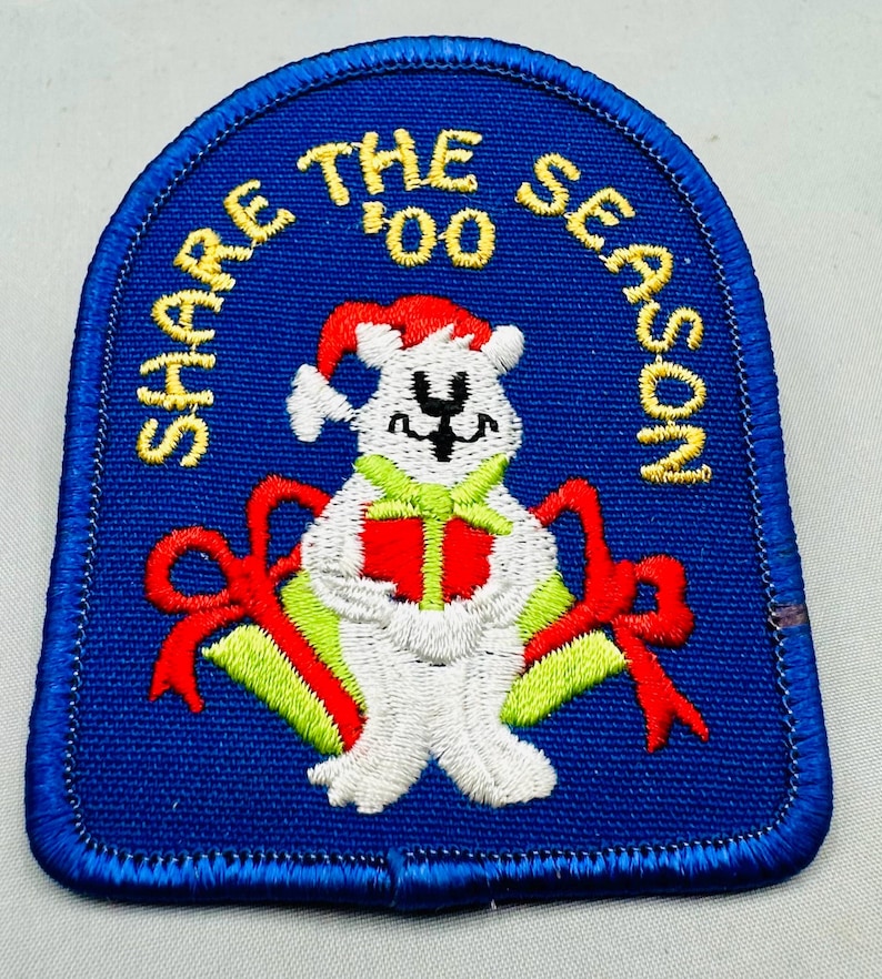 K&ouml;nnte beinhalten: Gestickter Aufn&auml;her mit blauem Hintergrund und einem wei&szlig;en B&auml;ren mit Weihnachtsm&uuml;tze. Der B&auml;r h&auml;lt ein gr&uuml;nes und rotes Geschenk. Der Text "SHARE THE SEASON '00" ist in Gelb gehalten. Rote Schleifen befinden sich auf beiden Seiten des B&auml;ren.
