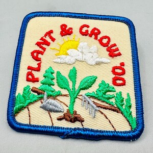 K&ouml;nnte beinhalten: Gestickter Aufn&auml;her mit den Worten "Plant & Grow" in Rot, mit einer gelben Sonne, wei&szlig;en Wolken, gr&uuml;nen B&auml;umen und einer sprie&szlig;enden Pflanze. Der Aufn&auml;her hat einen blauen Rand und einen beigen Hintergrund.