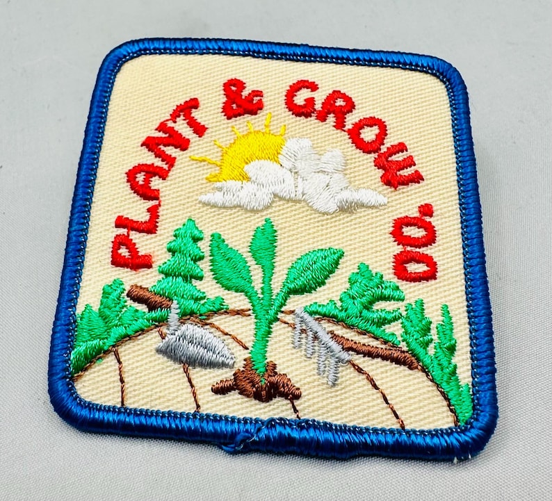 K&ouml;nnte beinhalten: Gestickter Aufn&auml;her mit den Worten "Plant & Grow" in Rot, mit einer gelben Sonne, wei&szlig;en Wolken, gr&uuml;nen B&auml;umen und einer sprie&szlig;enden Pflanze. Der Aufn&auml;her hat einen blauen Rand und einen beigen Hintergrund.