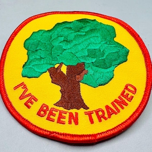 K&ouml;nnte beinhalten: Runder, gestickter Aufn&auml;her mit gelbem Hintergrund und rotem Rand. Der Aufn&auml;her zeigt einen gr&uuml;nen Baum mit braunem Stamm und dem roten Text "I'VE BEEN TRAINED".