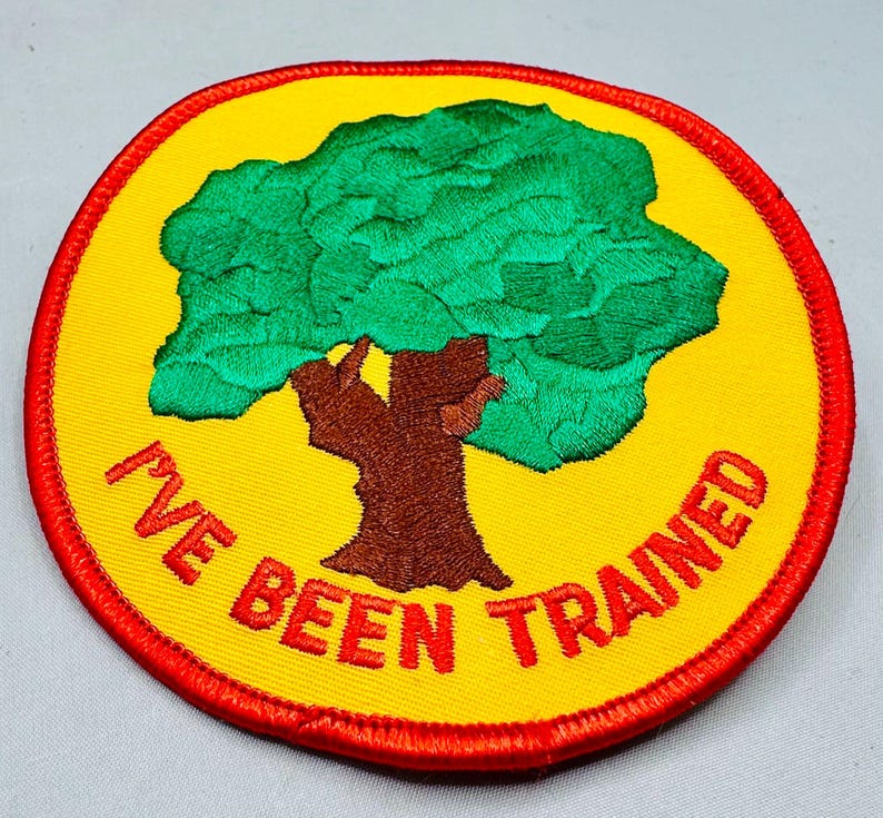 K&ouml;nnte beinhalten: Runder, gestickter Aufn&auml;her mit gelbem Hintergrund und rotem Rand. Der Aufn&auml;her zeigt einen gr&uuml;nen Baum mit braunem Stamm und dem roten Text "I'VE BEEN TRAINED".