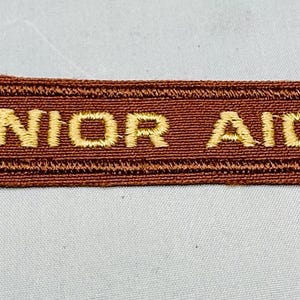 K&ouml;nnte beinhalten: Ein rechteckiger brauner Stoffaufn&auml;her mit den Worten "JUNIOR AIDE" in goldfarbenem Faden gestickt. Der Aufn&auml;her hat ein strukturiertes Aussehen und eine leicht abgewinkelte Kante auf der linken Seite.
