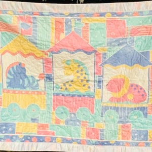 May include: A pastel-colored baby quilt featuring three train cars, each with a different animal: a blue zebra, a yellow giraffe, and a pink lion. The quilt has a white ruffled border and a patchwork design with squares and rectangles.