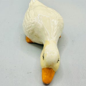 May include: A ceramic goose figurine with a glossy finish. The goose is primarily white with a yellow beak and feet. The figurine is in a downward-facing pose, suggesting it is pecking at the ground. The eyes are small and black.