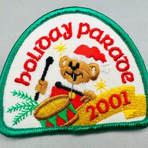 K&ouml;nnte beinhalten: Gestickter Aufn&auml;her mit wei&szlig;em Hintergrund, gr&uuml;nem Rand und den Worten "holiday parade" in Rot. Ein Teddyb&auml;r mit Weihnachtsm&uuml;tze spielt Trommel. Das Jahr "2001" steht auf einem gelben Banner.