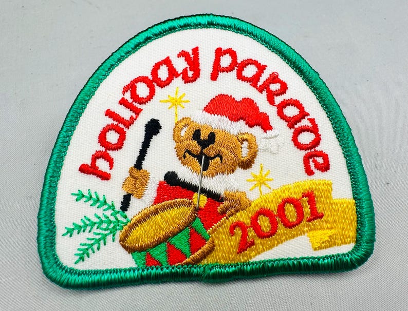 K&ouml;nnte beinhalten: Gestickter Aufn&auml;her mit wei&szlig;em Hintergrund, gr&uuml;nem Rand und den Worten "holiday parade" in Rot. Ein Teddyb&auml;r mit Weihnachtsm&uuml;tze spielt Trommel. Das Jahr "2001" steht auf einem gelben Banner.