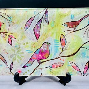 May include: Rectangular glass art piece featuring colorful birds perched on branches with leaves. The artwork has a light green and white background with pink, blue, and purple accents. The piece is displayed on a black stand.
