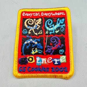 K&ouml;nnte beinhalten: Gestickter Aufn&auml;her mit rotem Hintergrund und gelbem Rand. Der Aufn&auml;her zeigt vier Quadrate mit farbenfrohen Designs und den Texten "Every Girl, Everywhere" und "GS Cookies 2002".