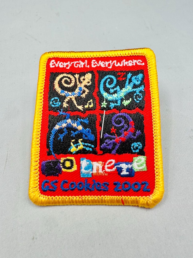 K&ouml;nnte beinhalten: Gestickter Aufn&auml;her mit rotem Hintergrund und gelbem Rand. Der Aufn&auml;her zeigt vier Quadrate mit farbenfrohen Designs und den Texten "Every Girl, Everywhere" und "GS Cookies 2002".