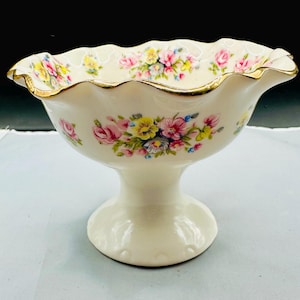 May include: Cream-colored pedestal bowl with a ruffled rim and gold trim. The bowl is decorated with clusters of pink, yellow, and blue flowers. A decorative piece for serving or display.