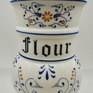 Puede incluir: Un bote de harina de cerámica blanca con tapa, adornado con motivos florales azules y naranjas. La palabra "flour" se muestra en negro. El bote tiene un borde azul y un adorno decorativo.