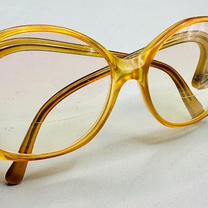 May include: Vintage oversized eyeglasses with a light amber-colored frame. The glasses have a distinctive, rounded shape and are held by a hand. The lenses are clear with a slight tint. A classic eyewear accessory.