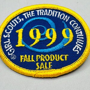 K&ouml;nnte beinhalten: Ein kreisf&ouml;rmiger Aufn&auml;her mit blauem Hintergrund und gelbem Rand. Der Text "GIRL SCOUTS, THE TRADITION CONTINUES" ist in Wei&szlig;. Das Jahr "1999" ist in Gelb, und der Text "FALL PRODUCT SALE" ist in Gelb.