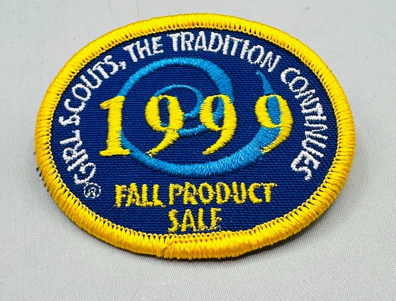K&ouml;nnte beinhalten: Ein kreisf&ouml;rmiger Aufn&auml;her mit blauem Hintergrund und gelbem Rand. Der Text "GIRL SCOUTS, THE TRADITION CONTINUES" ist in Wei&szlig;. Das Jahr "1999" ist in Gelb, und der Text "FALL PRODUCT SALE" ist in Gelb.