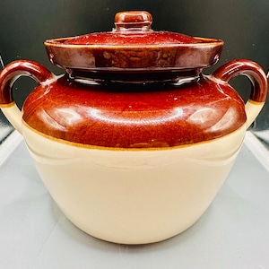May include: A vintage, two-toned ceramic crock pot with a lid. The crock pot is brown on top and white on the bottom, with two handles and a small knob on the lid. The crock pot is likely used for cooking or storage.