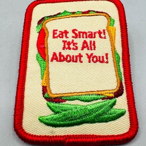K&ouml;nnte beinhalten: Rechteckiger Aufn&auml;her mit rotem Rand, der ein Sandwich-Design zeigt. Das Sandwich hat beiges Brot, rote Tomaten, gr&uuml;nen Salat und gelben K&auml;se. Der Text "Eat Smart! It's All About You!" ist rot.