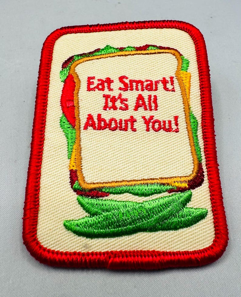 K&ouml;nnte beinhalten: Rechteckiger Aufn&auml;her mit rotem Rand, der ein Sandwich-Design zeigt. Das Sandwich hat beiges Brot, rote Tomaten, gr&uuml;nen Salat und gelben K&auml;se. Der Text "Eat Smart! It's All About You!" ist rot.