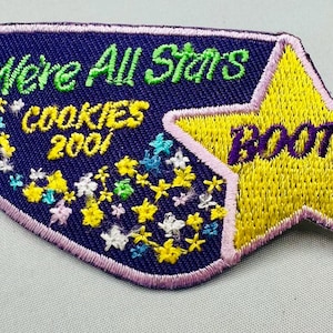 K&ouml;nnte beinhalten: Gestickter Aufn&auml;her mit marineblauem Hintergrund und gelbem Stern. Der Aufn&auml;her tr&auml;gt die Aufschrift "We're All Stars COOKIES 2001 BOOTH" in gr&uuml;ner, gelber und violetter Farbe. Der Aufn&auml;her ist rosa umrandet und mit gestickten Blumen versehen.