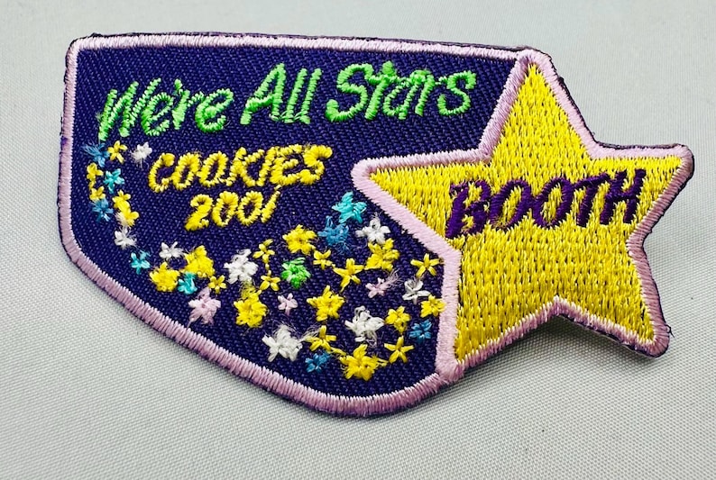 K&ouml;nnte beinhalten: Gestickter Aufn&auml;her mit marineblauem Hintergrund und gelbem Stern. Der Aufn&auml;her tr&auml;gt die Aufschrift "We're All Stars COOKIES 2001 BOOTH" in gr&uuml;ner, gelber und violetter Farbe. Der Aufn&auml;her ist rosa umrandet und mit gestickten Blumen versehen.