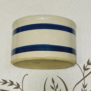 Puede incluir: Un tarro cilíndrico de gres con cuerpo color crema y dos franjas azules horizontales. El recipiente de estilo antiguo tiene una base ligeramente redondeada y probablemente se utiliza para almacenar alimentos o para decoración.