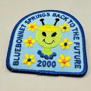 K&ouml;nnte beinhalten: Gestickter Aufn&auml;her mit hellblauem Hintergrund und dunkelblauem Rand. Der Aufn&auml;her zeigt ein l&auml;chelndes, gr&uuml;nes, au&szlig;erirdisch aussehendes Wesen mit gelben Blumen und dem Text "BLUEBONNET SPRINGS BACK TO THE FUTURE 2000".