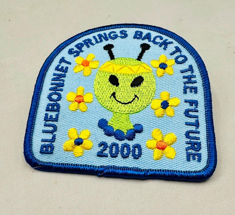K&ouml;nnte beinhalten: Gestickter Aufn&auml;her mit hellblauem Hintergrund und dunkelblauem Rand. Der Aufn&auml;her zeigt ein l&auml;chelndes, gr&uuml;nes, au&szlig;erirdisch aussehendes Wesen mit gelben Blumen und dem Text "BLUEBONNET SPRINGS BACK TO THE FUTURE 2000".