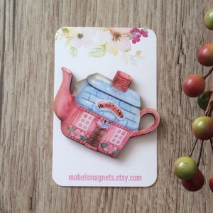 Könnte beinhalten: Ein dekorativer Magnet in Form eines Teekannenhauses. Die Teekanne ist rosa bemalt mit einem blauen Dach und einem roten Schornstein. Das Wort "HEARTLAND" steht auf einem Banner. Der Magnet befindet sich auf einer weißen Karte mit floralen Akzenten. Die Website mabelsmagnets.etsy.com ist auf der Karte aufgedruckt.