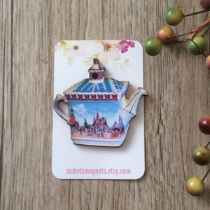 Könnte beinhalten: Ein Holz-Teekannen-Magnet mit einem blauen und weißen Design, das ein Cartoon-Bild einer Burg und Menschen zeigt. Der Magnet befindet sich auf einer weißen Karte mit dem Text "mabelsmagnets.etsy.com".