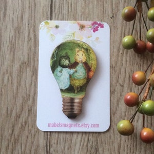 Peut inclure: Un aimant en bois en forme d'ampoule avec une illustration vintage de deux animaux à l'intérieur. L'aimant est sur une carte blanche avec des motifs floraux roses et le texte "mabelsmagnets.etsy.com".