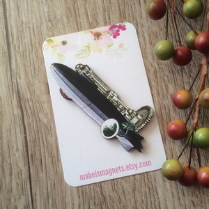 Pode incluir: Um broche de madeira em forma de dirigível steampunk com corpo preto, engrenagens prateadas e uma hélice verde. O broche está em um cartão branco com o texto "mabelsmagnets.etsy.com".