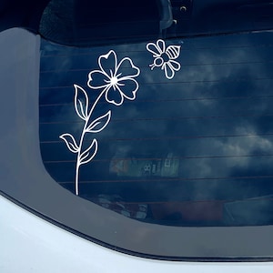 May include: White vinyl decal of a flower with a bee. The flower has a large bloom with multiple petals and a long stem with leaves. The bee is flying near the flower. The decal is designed for application on a car window.
