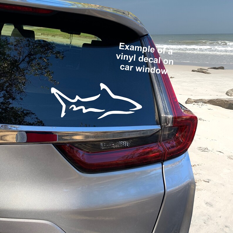 K&ouml;nnte beinhalten: Ein wei&szlig;er Hai-Silhouetten-Aufkleber aus Vinyl auf einem silbernen Autofenster. Der Aufkleber ist ein Beispiel f&uuml;r einen Autofensteraufkleber. Das Auto steht auf einem Sandstrand mit dem Meer im Hintergrund.
