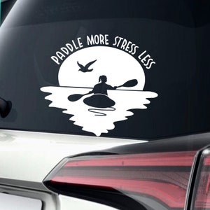 May include: White vinyl decal with a silhouette of a person kayaking on a lake with a bird flying overhead. The text "Paddle More Stress Less" is written in a circle around the image.