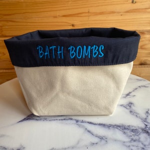 May include: A cream-coloured canvas bath bomb storage container with a navy blue fabric top.  The words 'BATH BOMBS' are embroidered in teal on the navy blue fabric. The container is shown on a marble surface.